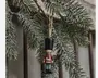 En lille hængende nøddeknækkerfigur i træ med høj sort hat, bordeaux jakke, lyseblå bukser og sorte støvler, ophængt i en snor blandt frostede granաչer.