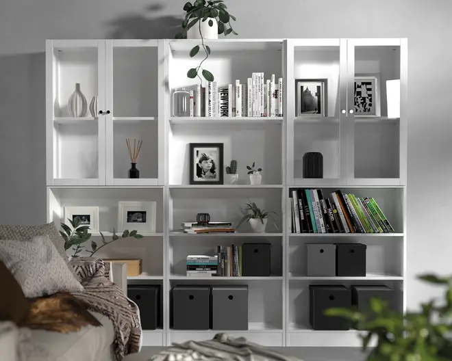 En stor, hvid reol med flere rum, hvor nogle har glaslåger med runde håndtag. Hylderne er organiseret med en blanding af bøger, indrammede fotografier, dekorative vaser, potteplanter og sorte opbevaringskasser. Udtrykket er moderne og ryddeligt med en kombination af åbne og lukkede opbevaringsmuligheder. Reolen står op ad en lysegrå væg, og øverste hylde har en grøn plante med hængende ranker.