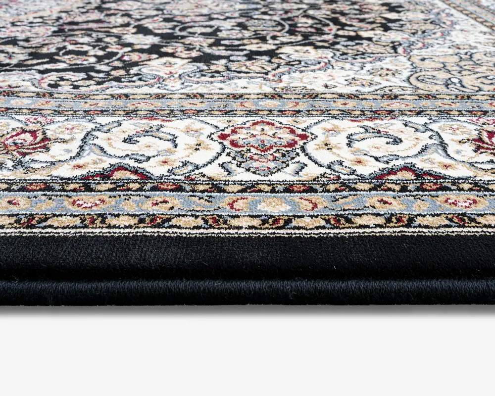 Nærbillede af et tykt, blødt tæppe med en detaljeret og udsmykket kant, der viser indviklede blomster- og snirklemønstre i nuancer af creme, rød, blå og sort. Tæppet har en tæt luv, hvilket giver en blød og polstret overflade. Farverne og designet antyder en klassisk, elegant stil, der passer til traditionelle indretninger.