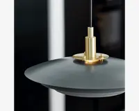 Nærbillede af en moderne pendellampe med en bred, mat sort cirkelformet skærm og et elegant, børstet guldfarvet metalbeslag øverst. Lampen hænger i en tynd sort ledning, og designet forener moderne elegance med minimalistisk stil.