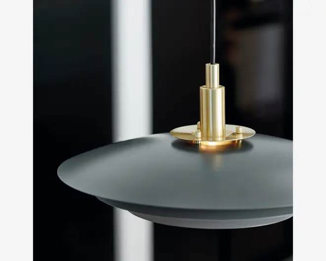 Nærbillede af en moderne pendellampe med en bred, mat sort cirkelformet skærm og et elegant, børstet guldfarvet metalbeslag øverst. Lampen hænger i en tynd sort ledning, og designet forener moderne elegance med minimalistisk stil.