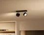 Sort loftmonteret lampe med smal rektangulær base og to justerbare cylindriske hoveder, hvor det ene peger nedad og lyser varmt, mens det andet er vinklet til siden i et beige rum.