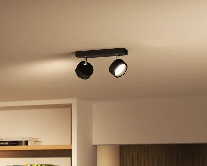 Sort loftmonteret lampe med smal rektangulær base og to justerbare cylindriske hoveder, hvor det ene peger nedad og lyser varmt, mens det andet er vinklet til siden i et beige rum.