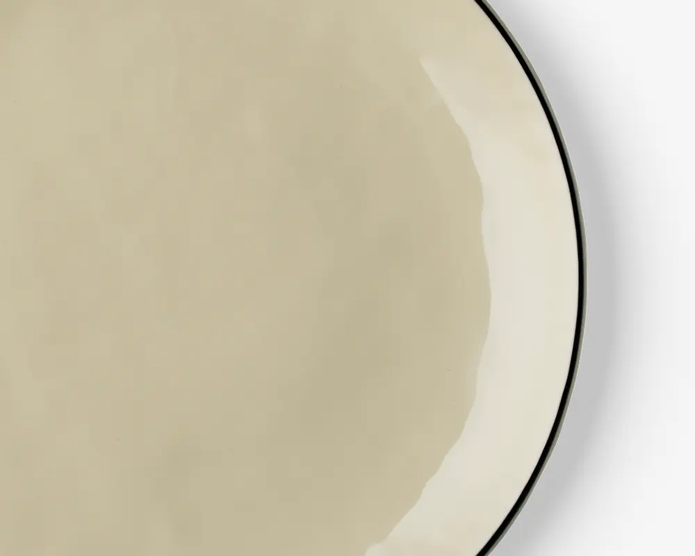 Et nærbillede af en rund, beige tallerken med en glat, blank overflade. Tallerkenen har en diskret, lidt mørkere kant og et minimalistisk design uden mønstre eller dekorationer. Overfladen virker jævn, og kanten er let buet.