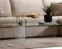 Ovalt sofabord fremstillet udelukkende af klart glas med et minimalistisk design, flad bordplade og to vinkelrette glasplader som ben. På bordet står dekorative genstande, herunder en sort rillet urtepotte med en grøn plante, en stak magasiner, et hvidt stearinlys i en lille holder og en lille vase med tørrede blomster. Det gennemsigtige materiale giver bordet et let og luftigt udtryk, der passer godt ind i moderne eller nutidige indretninger.