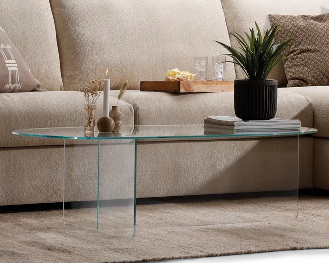 Ovalt sofabord fremstillet udelukkende af klart glas med et minimalistisk design, flad bordplade og to vinkelrette glasplader som ben. På bordet står dekorative genstande, herunder en sort rillet urtepotte med en grøn plante, en stak magasiner, et hvidt stearinlys i en lille holder og en lille vase med tørrede blomster. Det gennemsigtige materiale giver bordet et let og luftigt udtryk, der passer godt ind i moderne eller nutidige indretninger.