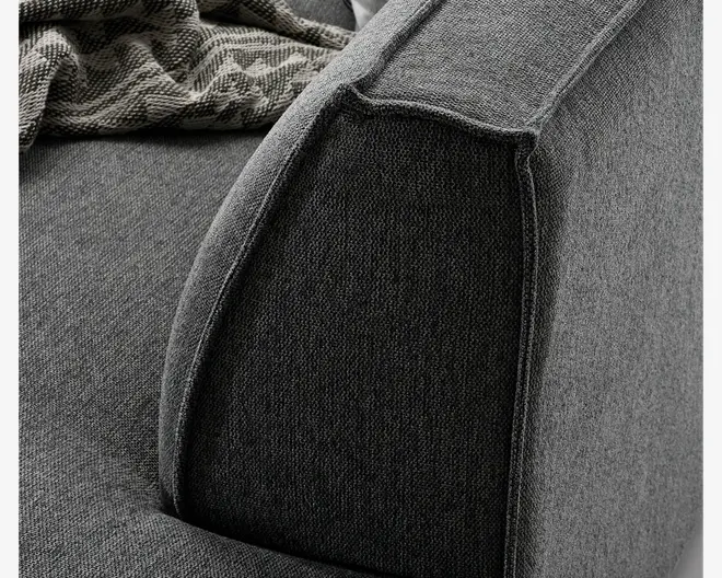 Nærbillede af en sofa-armlæn med tekstureret mørkegrå stofpolstring. Armlænet har en let afrundet form med synlige sømme, der markerer strukturen. Sædehynden og ryglænet er også dækket af samme stof, og et mønstret tæppe ligger over ryglænet og giver kontrast.
