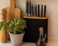 Et sæt på seks køkkenknive med sorte håndtag og sølvfarvede spidser, pænt arrangeret i en moderne knivblok i sort og træ. Blokken har et glat, rektangulært design med en trætop. Knivene varierer i størrelse og form, velegnet til forskellige køkkenopgaver. Knivblokken står på en bordplade ved siden af en potte med basilikum, en træpeberkværn, to skærebrætter i træ og en glasflaske med olivenolie med korkprop.