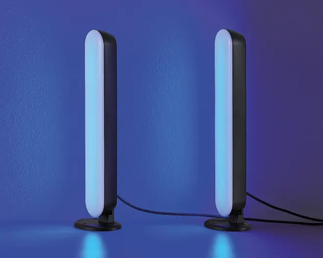 Et par slanke, lodrette LED-lysstænger med afrundede kanter, der udsender et blødt blåt skær. Hver lysstang er monteret på en rund sort fod og forbundet med synlige sorte strømkabler. Lysene står oprejst på en flad overflade op ad en blå væg, hvilket giver et moderne og minimalistisk udtryk.
