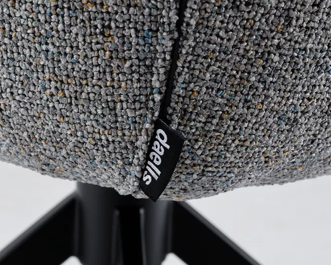 Nærbillede af et gråt tekstilbetræk med små blå og gule prikker, samt et sort stofmærke med hvid 'daells' branding. Betrækket er monteret på et sæde med sort metalbase og ben, som ses nederst i billedet.