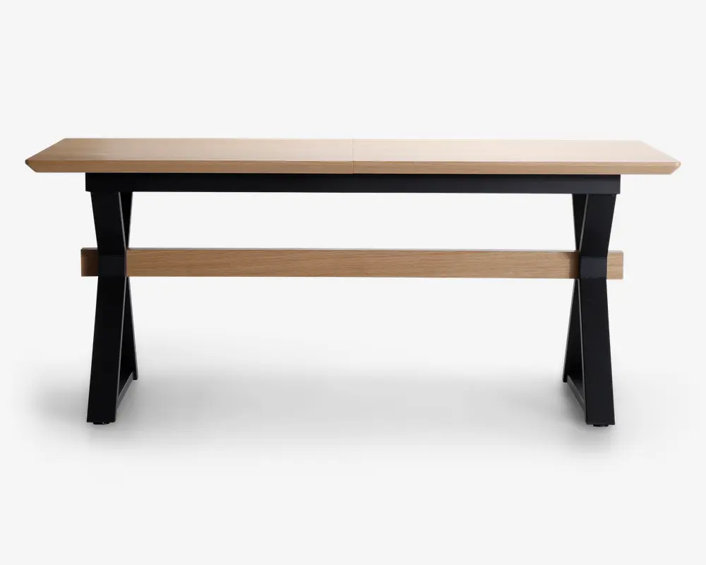 Rektangulært bord med en lys træbordplade og et solidt stel. Benene er skråtstillede og malet sorte, hvilket giver en moderne kontrast til det naturlige træ. En vandret træstang forbinder benene for ekstra stabilitet. Designet er minimalistisk og moderne, velegnet til spise- eller arbejdsbrug.