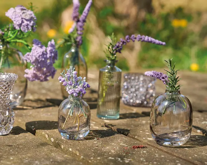 En samling små, klare glasvaser i forskellige former og teksturer, hver med et par stilke lilla og lavendelfarvede blomster. Vaserne inkluderer runde, riflede og facetslebne designs og er arrangeret på en rustik træoverflade udendørs. Glasset er gennemsigtigt, så stilke og vand indeni er synlige. Udtrykket er elegant og dekorativt, velegnet til at fremvise enkelte stilke eller små blomsterarrangementer.