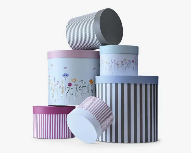 Sæt af cylindriske opbevaringsæsker med matchende låg i pastelblå, rosa og grå nuancer, med blomsterprint og lodrette stribemønstre i forskellige størrelser.