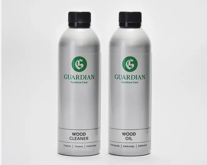 To høje, cylindriske sølvfarvede flasker med sorte skruelåg. Flasken til venstre er mærket 'Wood Cleaner', og flasken til højre er mærket 'Wood Oil'. Begge flasker har et grønt 'G'-logo og mærket 'Guardian Furniture Care'. Etiketterne er enkle og moderne med tydelig sort tekst på hvid baggrund nederst på hver flaske. Flaskerne ser ud til at være lavet af metal og er designet til nem hældning og opbevaring.