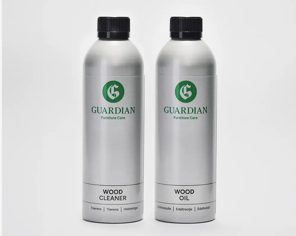 To høje, cylindriske sølvfarvede flasker med sorte skruelåg. Flasken til venstre er mærket 'Wood Cleaner', og flasken til højre er mærket 'Wood Oil'. Begge flasker har et grønt 'G'-logo og mærket 'Guardian Furniture Care'. Etiketterne er enkle og moderne med tydelig sort tekst på hvid baggrund nederst på hver flaske. Flaskerne ser ud til at være lavet af metal og er designet til nem hældning og opbevaring.