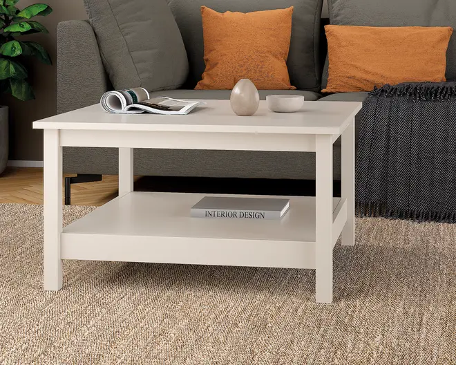 Rektangulært sofabord med en glat, lysefarvet overflade. Bordet har et robust design med lige ben og en ekstra hylde nederst til opbevaring, hvor der ligger en bog. Bordpladen er rummelig og viser et magasin, en dekorativ vase og en lille skål. Bordet står på et tekstureret tæppe foran en sofa og passer godt ind i moderne stueindretning.