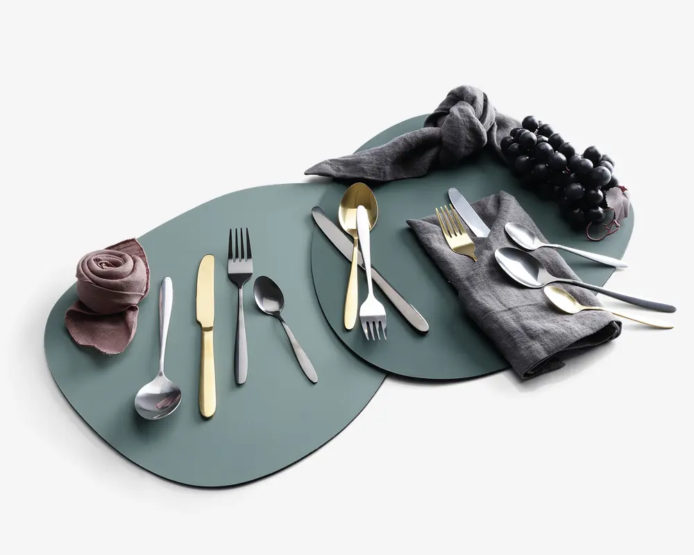 Et sæt moderne bestik præsenteret på tre overlappende, matte grønne dækkeservietter. Bestikket består af gafler, knive og skeer i en blanding af sølv- og guldfinish med slanke, minimalistiske håndtag. Opsætningen inkluderer sammenrullede og foldede stofservietter i afdæmpede farver samt en klase sorte druer som dekoration. Stilen er nutidig og elegant, velegnet til et stilfuldt bordopdækning.