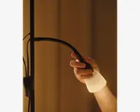 Et nærbillede af en hånd, der justerer en fleksibel lampe med en sort, bøjelig hals og en mat hvid lampeskærm. Lampens hals kan nemt bøjes, så lyset kan rettes i forskellige retninger. Lampen udsender et varmt, blødt lys, og baggrunden er svagt oplyst, hvilket skaber en hyggelig stemning.