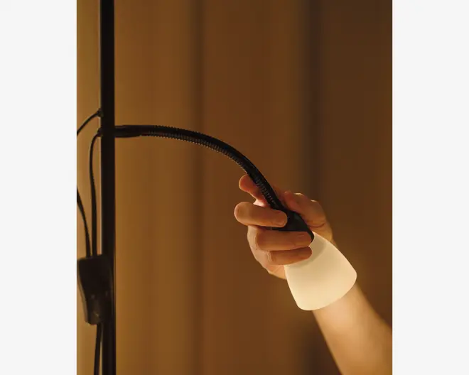Et nærbillede af en hånd, der justerer en fleksibel lampe med en sort, bøjelig hals og en mat hvid lampeskærm. Lampens hals kan nemt bøjes, så lyset kan rettes i forskellige retninger. Lampen udsender et varmt, blødt lys, og baggrunden er svagt oplyst, hvilket skaber en hyggelig stemning.