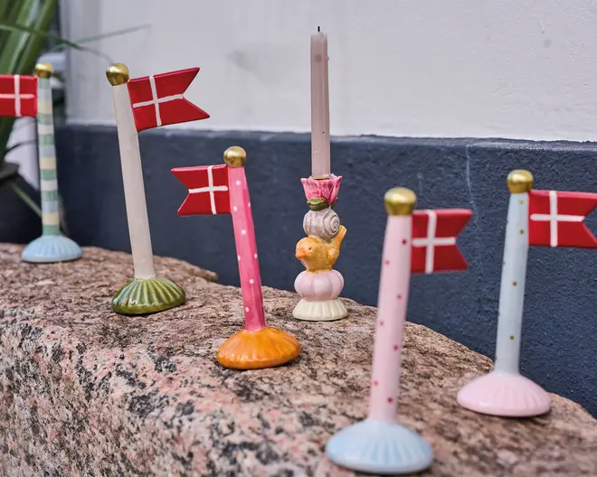 En samling dekorative flagholdere, hver med et lille dansk flag fastgjort til en farverig, rund fod. Fødderne har forskellige farver og mønstre, herunder striber, prikker og ensfarvede, alle med guldfarvede toppe. Én holder i midten er udformet som en lysestage med et fantasifuldt design med en snegl og en kylling, og holder et lyserødt stearinlys. Produkterne er udstillet på en stenoverflade op ad en blå og hvid væg.