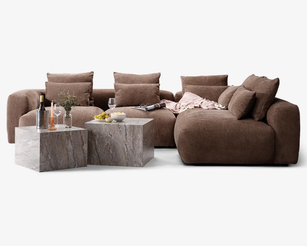 En modulopbygget sofa med blødt, dybbrunt stofbetræk og ekstra tykke ryg- og sædehynder. Opsætningen inkluderer en chaiselong-del og flere store, løse puder for ekstra komfort. Foran sofaen står to firkantede sofaborde med marmorlignende overflade i grå og hvide årer. Bordene er dekoreret med glas, en vinflaske, en vase med grønne grene og små skåle med snacks, hvilket skaber en hyggelig og indbydende stemning.