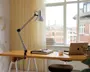 Justerbar skrivebordslampe med metalfinish, udstyret med en fjederbelastet arm og klemmebase, der fastgøres sikkert til kanten af et træskrivebord. Lampen har en konisk lampeskærm og kan placeres i forskellige vinkler for målrettet belysning. Designet er moderne og funktionelt, velegnet til arbejds- eller studiemiljøer.