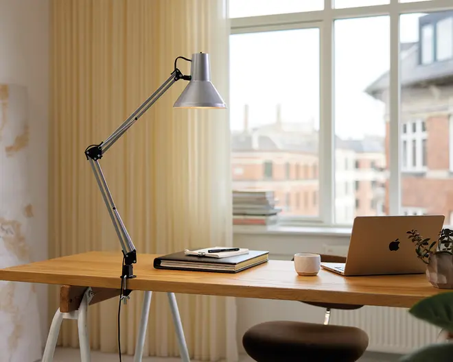 Justerbar skrivebordslampe med metalfinish, udstyret med en fjederbelastet arm og klemmebase, der fastgøres sikkert til kanten af et træskrivebord. Lampen har en konisk lampeskærm og kan placeres i forskellige vinkler for målrettet belysning. Designet er moderne og funktionelt, velegnet til arbejds- eller studiemiljøer.