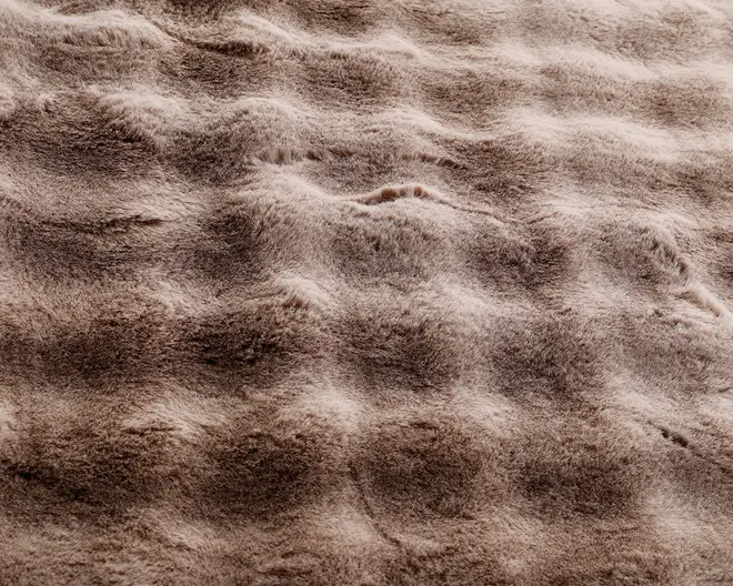 Nærbillede af en blød, plysset overflade med bølget, tekstureret mønster. Materialet fremstår tykt og luftigt med nuancer af lyse og mellembrune farver. Teksturen giver en følelse af dybde og komfort, hvilket antyder en hyggelig og luksuriøs fornemmelse.