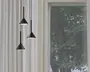 Tre moderne pendellamper med mat sorte, kegleformede skærme hænger i forskellige højder. Hver lampe har en slank, sort ledning og et minimalistisk design, der udsender et blødt, hvidt lys nedad. Baggrunden viser hvide gardiner og et vindue med udsigt til grønne træer udenfor.