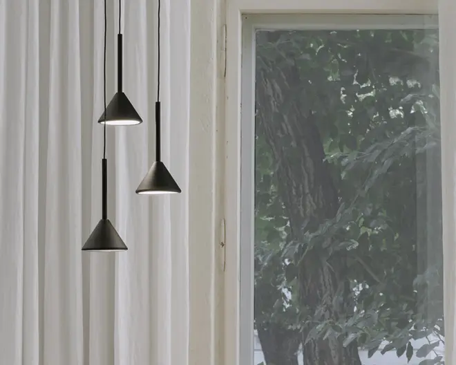 Tre moderne pendellamper med mat sorte, kegleformede skærme hænger i forskellige højder. Hver lampe har en slank, sort ledning og et minimalistisk design, der udsender et blødt, hvidt lys nedad. Baggrunden viser hvide gardiner og et vindue med udsigt til grønne træer udenfor.