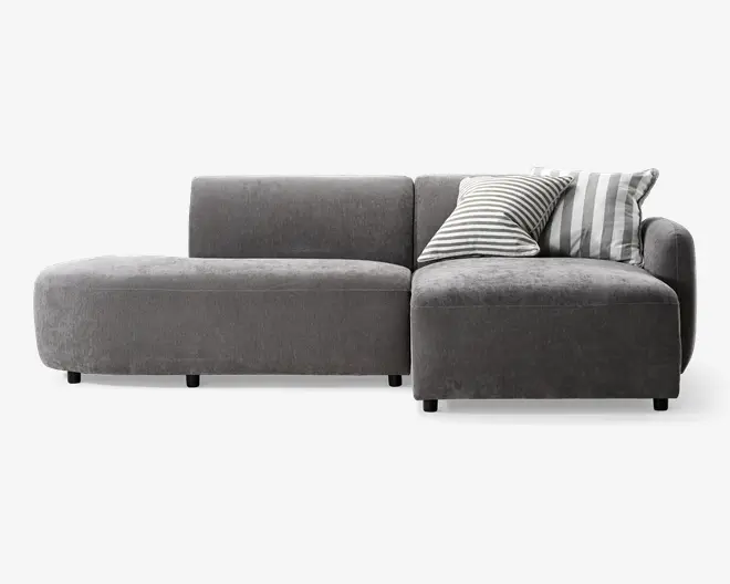 Moderne hjørnesofa med lav profil, betrukket med blødt, mørkegråt stof. Sofaen har en venstrevendt chaiselong og et højrevendt sæde med lige armlæn. Der medfølger to pyntepuder: én med smalle sorte og hvide striber og én med brede grå og hvide striber. Sofaen har rene linjer og et minimalistisk udtryk, understøttet af korte, solide ben.