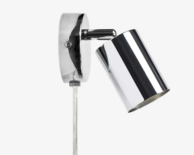 En vægmonteret lampe med blank kromfinish. Lampen har en cylindrisk skærm, der kan vinkles for justerbar belysning. Basen er oval og også i krom, med en synlig skrue til montering og en gennemsigtig ledning, der går nedad. Designet er moderne og stilrent, velegnet til nutidige indretninger.
