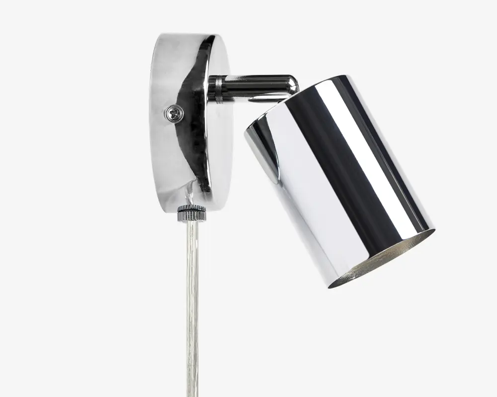 En vægmonteret lampe med blank kromfinish. Lampen har en cylindrisk skærm, der kan vinkles for justerbar belysning. Basen er oval og også i krom, med en synlig skrue til montering og en gennemsigtig ledning, der går nedad. Designet er moderne og stilrent, velegnet til nutidige indretninger.