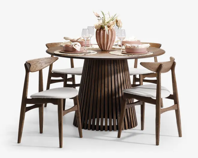 Rundt spisebord med en sokkel af lodrette trælameller og matchende træstole med buede ryglæn og hvide polstrede sæder. Bordpladen er dækket med flettede dækkeservietter, lyserøde servietter, glas og en dekorativ vase med lyse blomster, hvilket giver et elegant og indbydende spiseområde.