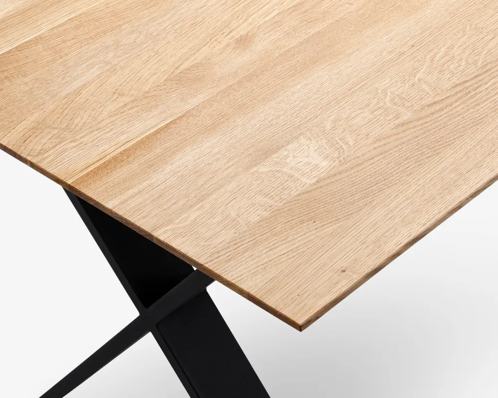 Nærbillede af en bordplade med en lys, naturlig træstruktur, der har en glat, flad overflade og let affaset kant. Bordpladen understøttes af en solid sort metalramme med krydsende ben, hvilket giver et moderne og minimalistisk udtryk.