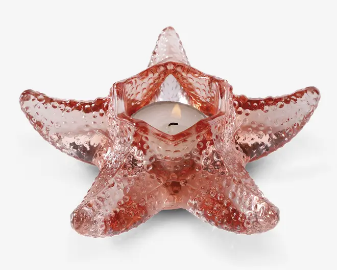 Et dekorativt fyrfadsstage formet som en søstjerne, lavet af lyserødt glas med en nubret overflade, der giver et realistisk udtryk. Stagen har et centralt hulrum, der passer til et lille fyrfadslys, som er tændt på billedet. Glasset er gennemsigtigt og lader lyset skinne blødt igennem.