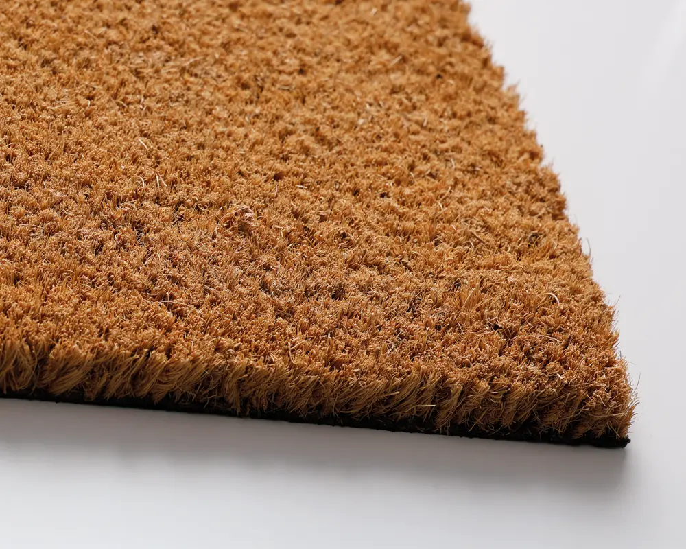 Nærbillede af en tyk, rektangulær måtte med en grov, tekstureret overflade lavet af naturlige brune fibre. Måtten har en tæt, stiv luv, der giver en ru fornemmelse, ideel til at skrabe snavs af sko. Kanterne er skarpt afskåret, og bagsiden virker solid, hvilket sikrer holdbarhed og stabilitet på gulvet.