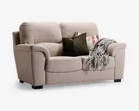 En to-personers sofa betrukket med lysebeige stof og bløde, polstrede ryglæn og armlæn. Sofaen har to sædehynder og står på lave, mørke træben. Den er pyntet med to dekorative puder – én ensfarvet mørkegrøn og én med røde og beige striber – samt et tekstureret, frynset gråt tæppe, der er lagt over det ene armlæn.