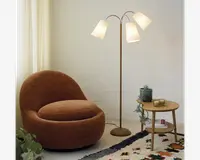 En hyggelig, rund lænestol betrukket med blødt, rustfarvet stof og en tyk, blød siddepude. Stolen har et lavt design og et moderne, indbydende udtryk. Ved siden af står en slank gulvlampe i gylden finish med tre buede arme, hver med en hvid, kegleformet lampeskærm. Et lille sidebord i lyst træ med skrå ben har en blå vase med grønne planter, to stearinlys og en bog på den nederste hylde. Rummet fuldendes af et cremefarvet gardin og et mønstret tæppe med farverige detaljer.
