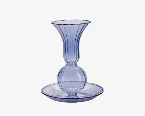 Gennemsigtigt lys lilla glas med en bred, riflet top, en rund midterdel og en bred, lav bund. Designet har lodrette riller langs kroppen, hvilket giver et elegant og dekorativt udtryk. Glasset har en glat og blank overflade.
