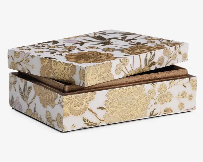 Rektangulær æske med hængslet låg, dekoreret med et hvidt baggrund og indviklede guldfarvede blomster-mønstre. Blomsterne og bladene er præget, hvilket giver overfladen en tekstureret og luksuriøs fornemmelse. Indersiden ser ud til at være foret med en metallisk guldfinish, hvilket bidrager til det elegante og dekorative udtryk.