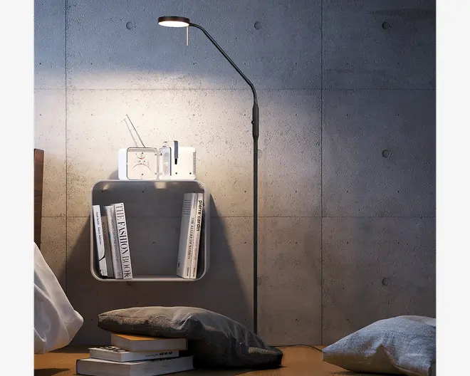 En slank, moderne gulvlampe med et minimalistisk design, der har en tynd, sort metalstang og en justerbar arm med et lille, rundt LED-lyshoved. Lampen giver fokuseret, nedadgående belysning, ideel til læsning eller arbejdslys. Basen er diskret, hvilket gør det nemt at placere lampen i små rum. Lampen er vist ved siden af en vægmonteret hylde med bøger og en lille radio, hvilket understreger dens egnethed til moderne indretning.