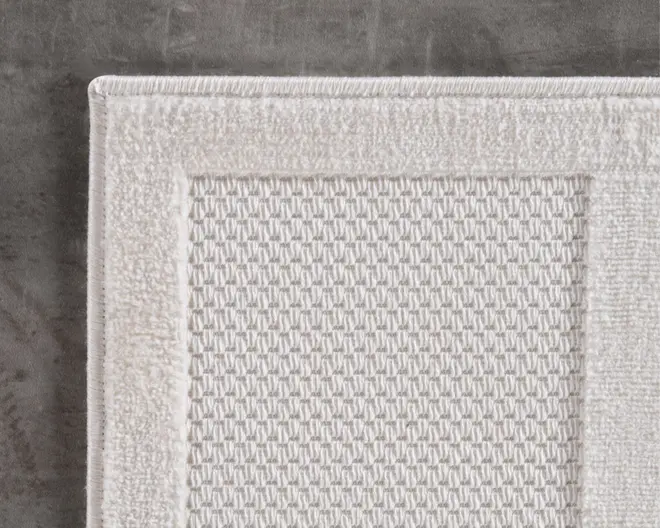 Nærbillede af en hvid tekstilgenstand med et tekstureret, vaffellignende mønster i midten og en bred, glat kant. Kanterne er pænt afsluttet med forstærket syning, hvilket giver et rent og holdbart udseende. Stoffet ser tykt og absorberende ud, velegnet til brug, hvor komfort og holdbarhed er vigtig.
