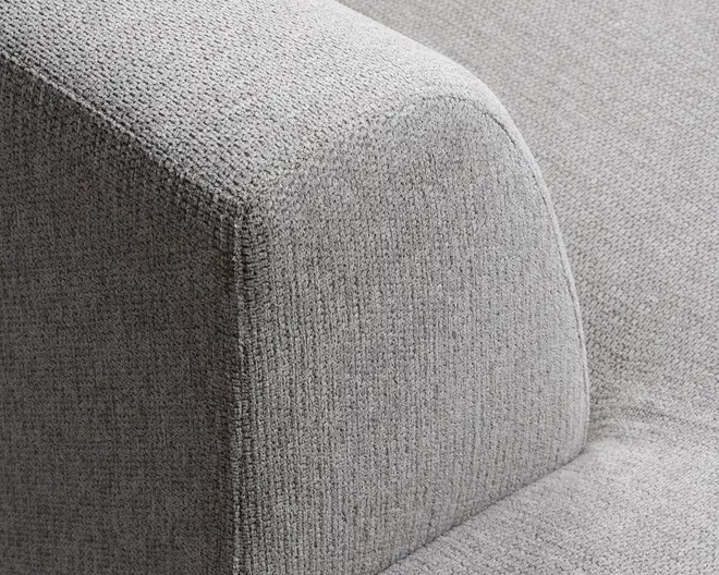 Nærbillede af et sofa-armlæn betrukket med lysegråt, tekstureret stof. Stoffet har et diskret vævet mønster, og armlænet har rene, moderne linjer med bløde, afrundede kanter. Det samlede udtryk er minimalistisk og moderne.