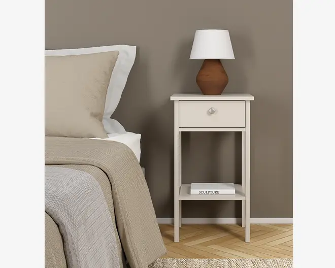 Dette er et kompakt, rektangulært natbord med en glat, lysebeige finish. Det har en enkelt skuffe med et rundt metalgreb for nem åbning samt en åben hylde nederst til ekstra opbevaring eller udstilling, her vist med en bog. Natbordet står på fire lige ben, hvilket giver stabilitet og et moderne, minimalistisk udtryk. Toppladen er flad og rummelig nok til at rumme genstande som en lampe eller dekoration.