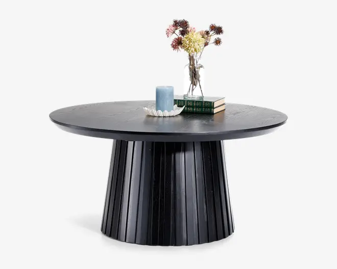 Rundt bord med en bred, rillet sokkel og en glat, mørk træfinish. Bordpladen er pyntet med en lille vase med tørrede blomster, et blåt lys på et dekorativt fad og to stablede bøger, hvilket giver et moderne og elegant udtryk.