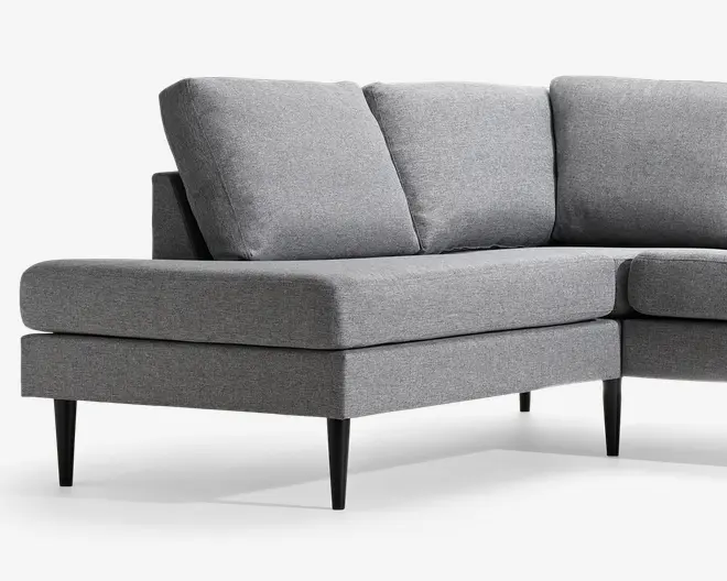 Lysegrå polstret hjørnesofa med bred chaiselong-lignende siddeplads, tre løse ryghynder og slanke sorte koniske ben i et rent, moderne design.