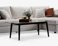 Rektangulært sofabord med en elegant, mørk glasplade og fire skråtstillede sorte metalben. Bordpladen er glat og reflekterende, og bordet er dekoreret med en træbakke, der indeholder et krus, et glas og en lille vase med grønne blade.