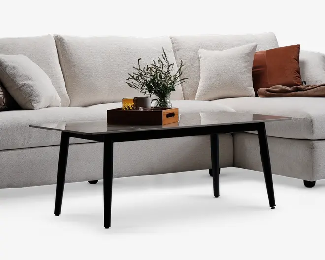 Rektangulært sofabord med en elegant, mørk glasplade og fire skråtstillede sorte metalben. Bordpladen er glat og reflekterende, og bordet er dekoreret med en træbakke, der indeholder et krus, et glas og en lille vase med grønne blade.