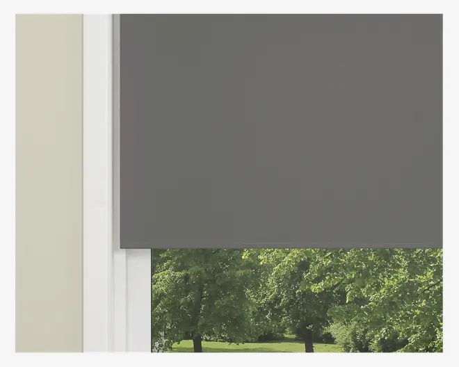 Et nærbillede af et vindue med en mørkegrå rullegardin, der er delvist rullet ned. Gardinet har et glat, ensfarvet stof og lige kanter. Vinduesrammen er hvid, og væggen ved siden af er lysebeige. Gennem den nederste, åbne del af vinduet ses grønne træer og græs udenfor, hvilket indikerer et naturligt udendørsmiljø.